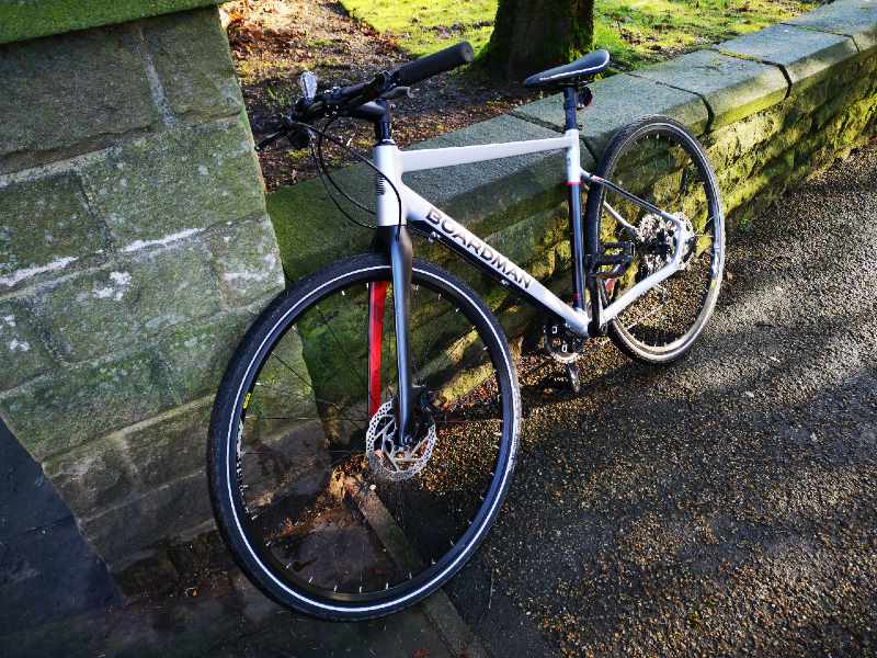 A silver boardman hybrid bike for cycling on roads and cross country too leaning against a wall in a park-hybrid bike-ss